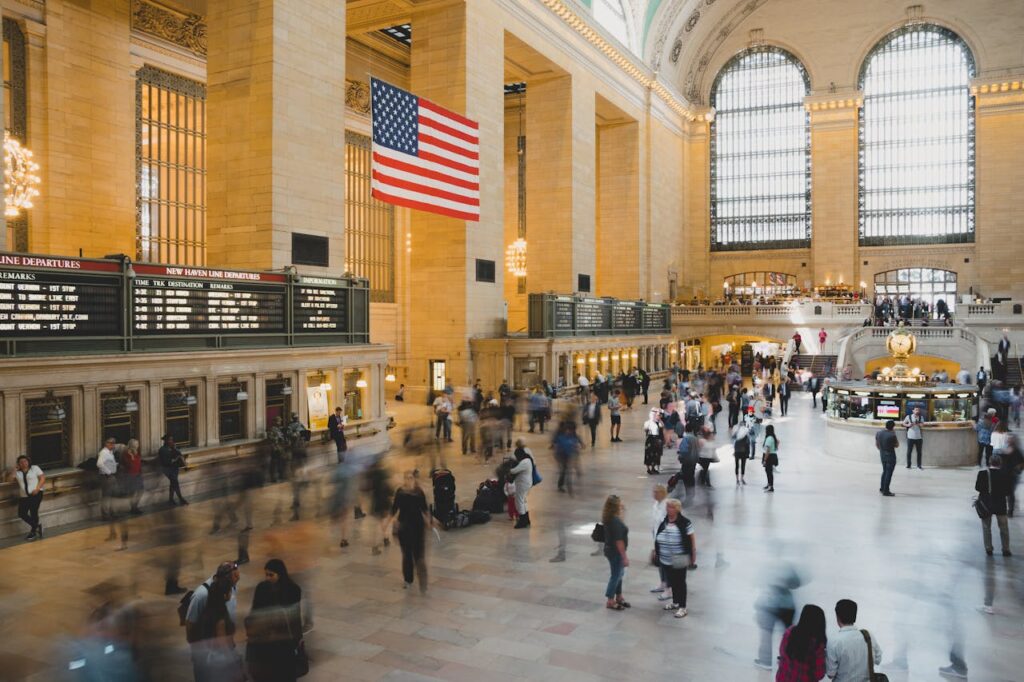 Crowd moves through historic Grand Central Terminal, New York City, under the American flag.